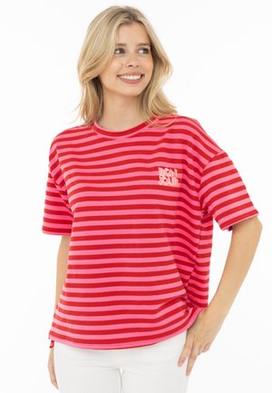 Blonde Frau lächelt, trägt ein rot-rosa horizontal gestreiftes T-Shirt mit dem Text "BON JOUR", kombiniert mit weißen Hosen, Hand auf der Hüfte.