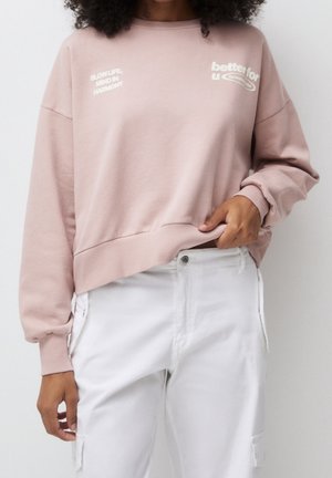 Person iført en lyserød sweatshirt med hvid tekst og hvide bukser, der holder i kanten af sweatshirtet med den ene hånd.