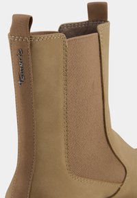 Bottine beige avec un corps en cuir lisse et un panneau élastique texturé sur le côté. Présente un design cousu et une languette à l'arrière.