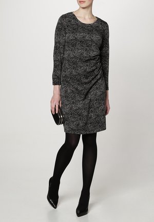 Robe noire et blanche avec des manches trois-quarts, un col rond et des détails froncés sur le côté. Portée avec des collants noirs et une petite pochette ronde.