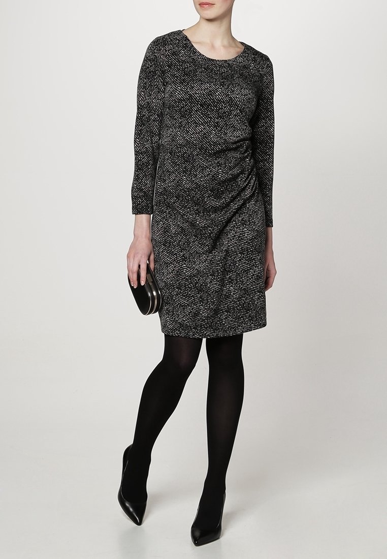Robe noire et blanche avec des manches trois-quarts, un col rond et des détails froncés sur le côté. Portée avec des collants noirs et une petite pochette ronde.