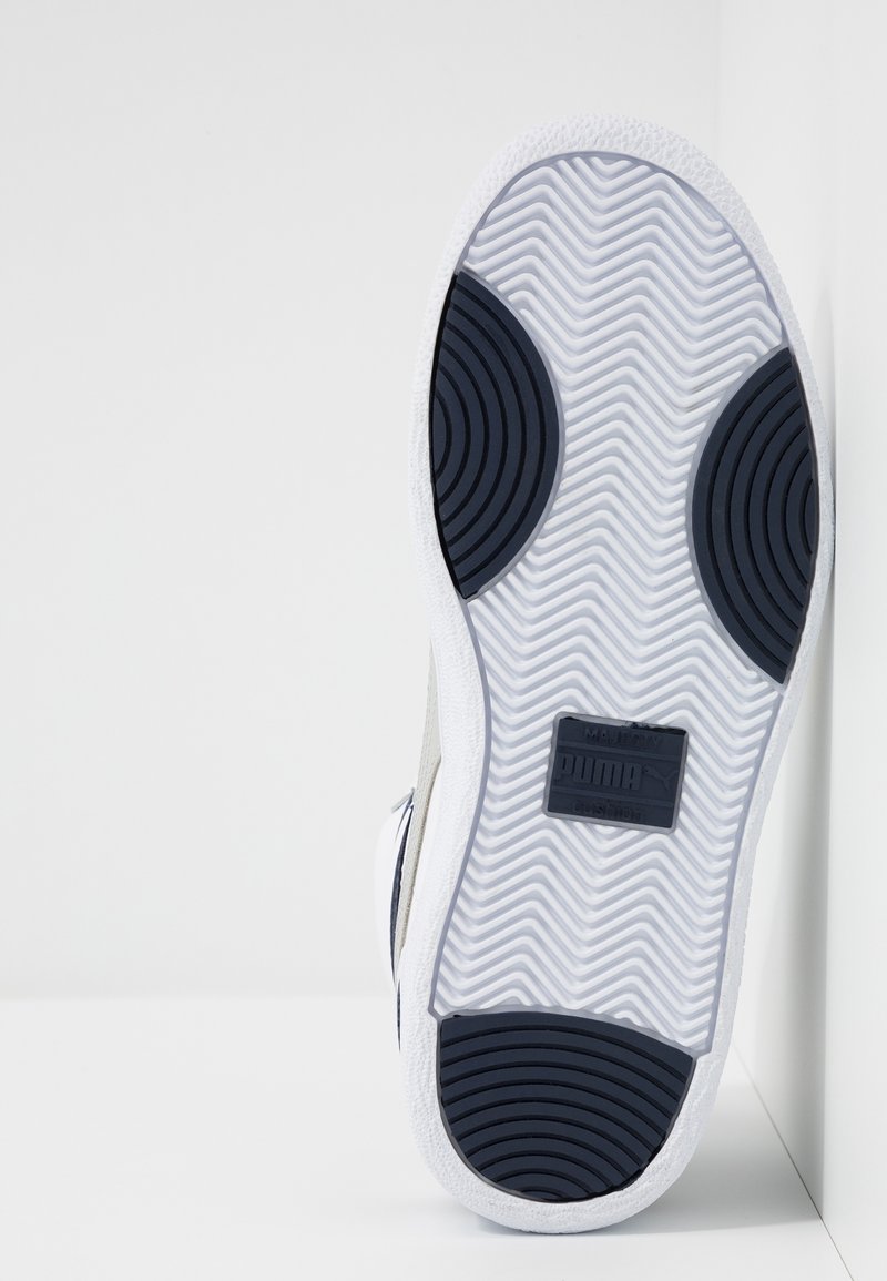 Semelle d'une basket blanche avec des motifs de grip circulaires noirs et une bande de roulement en zigzag, debout contre un mur d'angle blanc.
