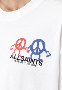 Símbolos de paz rojos y azules de personajes cogidos de la mano sobre el texto "ALLSAINTS" y "MENSAJE A UN FUTURO" en una camiseta de algodón blanca.