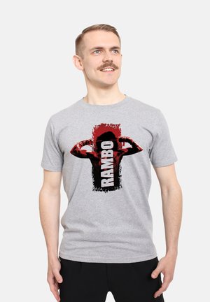 Grijze katoenen t-shirt met een opvallende zwart-rode graphic van een flexende figuur met de tekst "RAMBO" in vette letters.