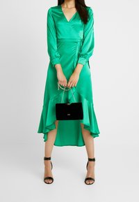 Abito avvolgente verde in satin con maniche lunghe a sbuffo e orlo asimmetrico, abbinato a una borsa nera in velluto e sandali neri con tacco.