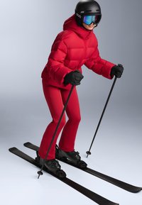 Piros puffadt dzseki és testhezálló piros nadrág fekete síbakancsokkal és sílécekkel. A fekete kesztyű és a sötétített szemüveges sisak teszi teljessé a ruházatot.