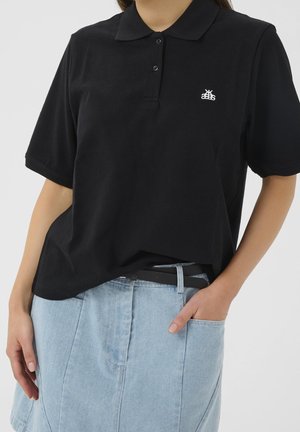 Woman wearing a black short-sleeve polo shirt with small white logo, tucked slightly into light blue denim skirt with black belt.