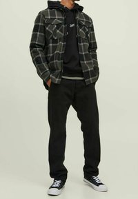 Giacca camicia a quadri neri e grigi con due tasche sul petto sopra un hoodie nero, abbinata a pantaloni neri e sneakers bianche e nere.