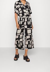 Mono con estampado en blanco y negro, con mangas cortas, cierre de botones, cintura con cordón y diseño de pierna ancha. Combinado con sandalias negras gruesas.
