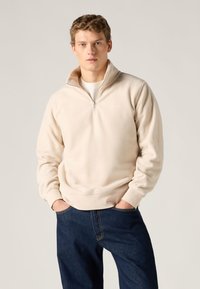 Ελαφρύ μπεζ φλις φούτερ με κολάρο μισού ζιπ και ριμπ τελείωμα. Φορεμένο με σκουρόχρωμο τζιν, με μπροστινές τσέπες και χαλαρή γραμμή.