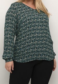 Blouse à manches longues en teal et crème à motif floral, avec un col en V, en tissu doux et coupe décontractée, associée à un pantalon noir.