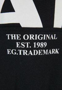 Fekete anyag fehér szöveggel: "AZ EREDETI EST. 1989 EG. VÉDJEGY." Enyhe textúrájú felület; tiszta, modern tipográfia.