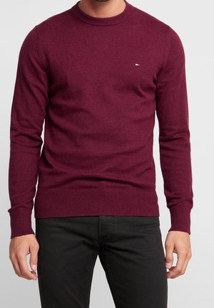 Homme portant un pull ajusté à manches longues couleur bordeaux avec un petit logo sur la poitrine gauche et un pantalon noir.