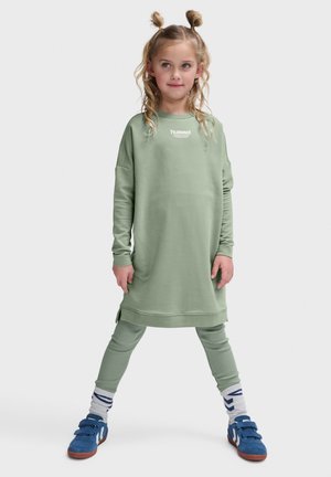 Lange groene sweatshirt jurk met verlaagde schouders en zijsplitten, gecombineerd met bijpassende groene legging en blauwe sneakers met klittenbandsluitingen.