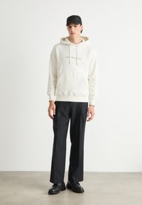 Sudadera crema con cordones y un bolsillo frontal, que presenta el logo "Calvin Klein Jeans". Combinada con pantalones negros a medida y zapatos negros.