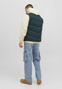 Padded navy blue vest with chevron stitching, worn over a beige hoodie, paired with light blue cargo jeans and brown boots.