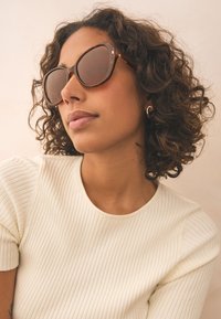 Solglasögon med sköldpaddsmönster och bruna linser, med en bred cat-eye-form. Kvinnan bär en vit ribbad topp med korta ärmar och rundad halsringning.