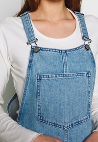 Vaaleansiniset farkkutakit, joissa on säädettävät metalliklipsit, etutasku ja hieman haalistunut pinta. Puettuna valkoisen pitkähihasen paidan päällä.