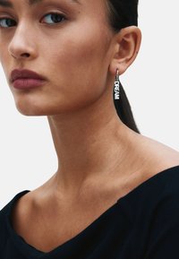 Sidabriniai lankelio auskarai su žodžiu „DREAM“ didžiosiomis raidėmis. Dėvimi modelio su tamsiais plaukais, vilkinčio juodą viršutinę dalį ir neutraliu fonu.