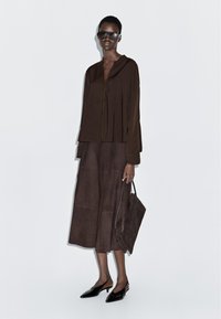 Brązowa, teksturowana bluzka z wiązaniem z przodu i szerokimi rękawami, zestawiona z brązową zamszową spódnicą. Czarne czółenka na szpilce z paskiem z tyłu oraz dopasowana torba typu tote.