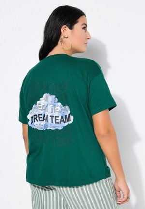 Frau mit langen dunklen Haaren, die große Creolen-Ohrringe trägt, ein grünes T-Shirt mit dem Schriftzug „DREAM TEAM“ und einer Wolkengrafik sowie grüne gestreifte Hose.