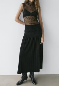 Woman wearing a sheer black sleeveless top, black pleated midi skirt, and black pointed-toe ankle boots, standing against a plain wall.