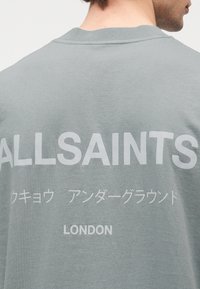 Γκρι βαμβακερό T-shirt με μεγάλο λευκό κείμενο στην πλάτη: "ALLSAINTS", ιαπωνικούς χαρακτήρες και "LONDON" στο κάτω μέρος.