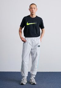 Černé tričko s krátkým rukávem Nike se zeleným logem, spárované se světle šedými tepláky s elastickým pasem a bočním potiskem a s manžetami na nohavicích.