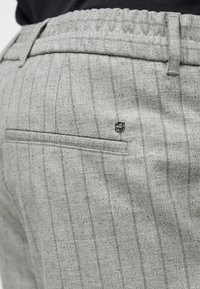 Siva, teksturirana tkanina z drobnimi črtami in zadnjim žepom. V bližini žepa je majhen kovinski logotip. Elastičen pas za udobje.