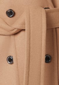 Beżowy wełniany płaszcz o teksturowanym wykończeniu, z czarnymi guzikami i związanym paskiem. Zbliżenie na zapięcie i szczegóły paska.