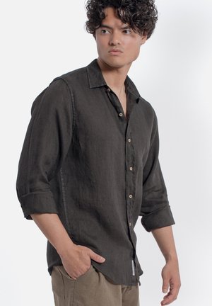 Giovane uomo che indossa una camicia grigio scuro con bottoni e maniche rimboccate e pantaloni beige, in piedi con una mano in tasca, guardando di lato.