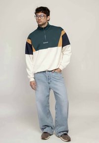 Ung mann står med den ene hånden i lomma, iført fargeblokkert half-zip genser, lyseblå vide jeans, joggesko og briller.