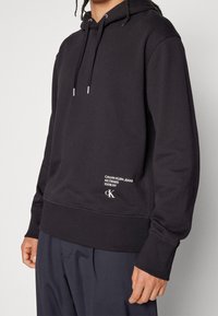 Osoba stojící před jednoduchým pozadím, oblečená v černé mikině Calvin Klein s bílým logem na přední kapse a tmavých kalhotách.