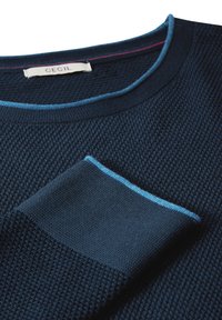 Maglione blu navy con design testurizzato, dettagli azzurri chiari lungo il girocollo e una tasca coordinata. Etichetta del brand visibile.