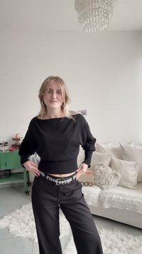 Svart kort tröja med vid halsringning och ribbad nederkant, kombinerad med svarta byxor med vit märkeslinning. Texturerad bakgrund.