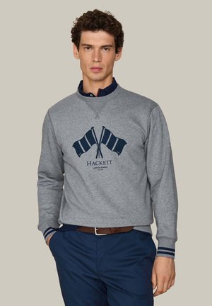 Junger Mann mit lockigem Haar trägt ein graues Sweatshirt mit dunkelblauen gekreuzten Flaggen und Text "Hackett London Rowing Club,", dunkelblaue Hose und einen braunen Gürtel.