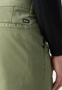 Pantaloni di stoffa verdi con una tasca posteriore che presenta un'etichetta nera "BOSS". Texture liscia e design sartoriale con una silhouette slim fit.