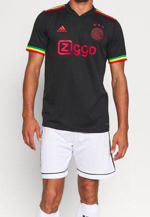 Homme portant un maillot de football noir de l'Ajax avec des bordures rouge, jaune et verte et un short blanc sur fond uni.