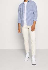 Muž oblečený v modro-bílém pruhovaném tričku přes bílé tričko, smetanových kalhotách a bílých teniskách Nike, stojící proti jednobarevnému neutrálnímu pozadí.