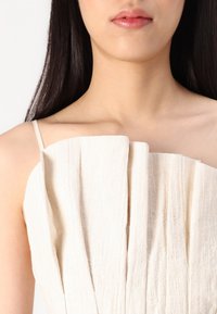 Robe crémeuse avec texture, au design plissé et fines bretelles, présentant un décolleté lisse et un subtil motif à lignes verticales.