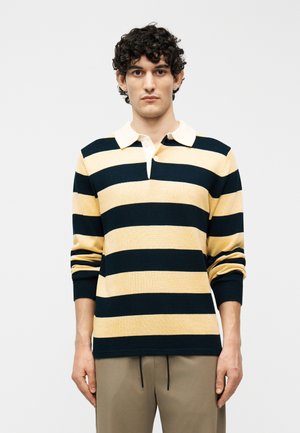 Jeune homme aux cheveux bouclés portant un maillot de rugby à manches longues avec de larges rayures horizontales noires et beiges, ainsi qu'un pantalon beige.