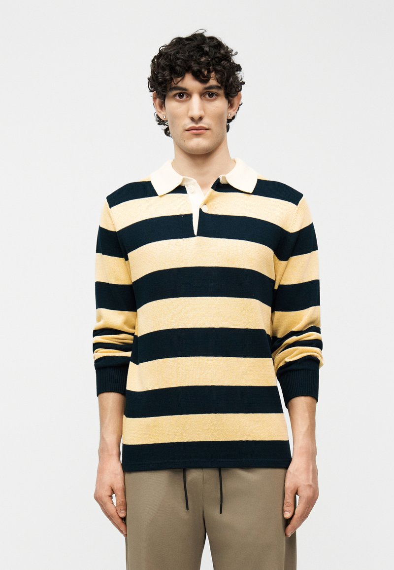 Jeune homme aux cheveux bouclés portant un maillot de rugby à manches longues avec de larges rayures horizontales noires et beiges, ainsi qu'un pantalon beige.