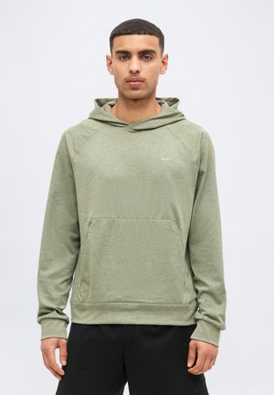 Young man with short hair wearing a light green hooded sweatshirt and black pants against a plain light background.