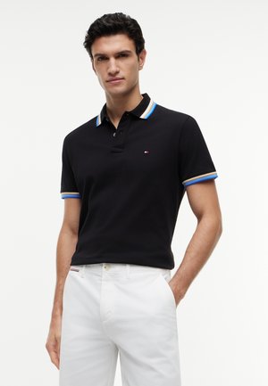 Homme portant un polo noir avec un col rayé bleu et jaune, et un pantalon blanc, debout avec une main dans la poche.