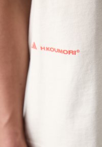 Vit bomull t-shirt med en mjuk textur som har en röd logotyp på vänster sida som står "H.KOUMORI®." Korta ärmar, avslappnad passform.