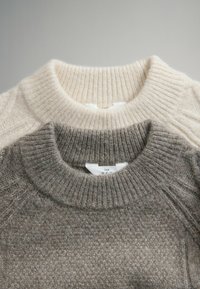 Twee truien met geribde halslijnen, één beige en één grijs. Beide zijn gemaakt van zacht gebreid materiaal en hebben een subtiele textuur. Labels zichtbaar.