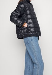 Black puffer jacket with a shiny finish, horizontal quilted texture, high collar, and brown cuff accents, paired with light blue jeans.