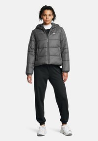 Grau gesteppte Daunenjacke mit Kapuze, Frontreißverschluss und horizontalem Steppmuster. Getragen mit schwarzen Jogginghosen und weißen Sportschuhen.