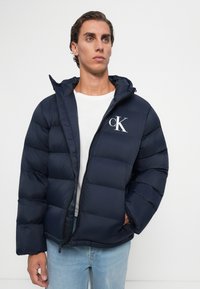 Jonge man in een marineblauwe pufferjack met het "cK" logo op de borst, een wit T-shirt en lichtblauwe jeans tegen een effen witte achtergrond.
