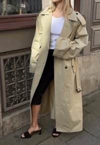 Light beige trench coat with a belted waist, large lapels, and black buttons, paired with black cropped pants and dark sandals.